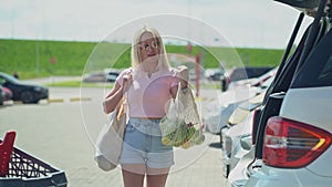 Young blonde smiling woman with shopping eco bags stay close to her opened car trunk on a parking