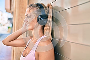 Young blonde sporty girl using headphones leaning on the wall at street of city