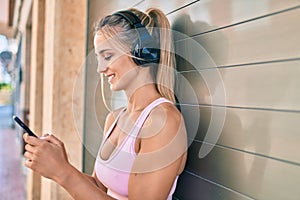 Young blonde sporty girl using smartphone and headphones leaning on the wall at street of city