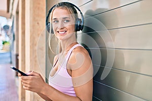 Young blonde sporty girl using smartphone and headphones leaning on the wall at street of city