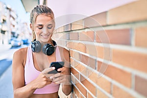 Young blonde sporty girl using smartphone and headphones leaning on the wall at street of city