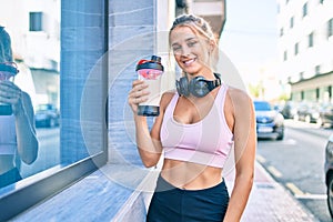 Young blonde sporty girl wearing headphones drinking healthy protein smoothie at street of city
