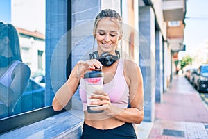 Young blonde sporty girl wearing headphones drinking healthy protein smoothie at street of city