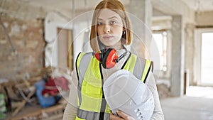 Young blonde woman architect standing with relaxed expression holding hardhat at construction site