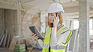 Young blonde woman architect talking on smartphone looking touchpad at construction site