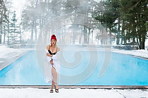 Young blonde woman in bathrobe and red hut near outdoor swimming pool at winter