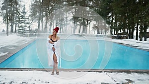 Young blonde woman in bathrobe and red hut near outdoor swimming pool at winter