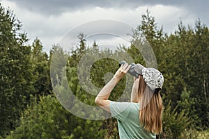 Young blonde woman bird watcher in cap and blue t-shirt looking through binoculars at cloudy sky in summer forest ornithological