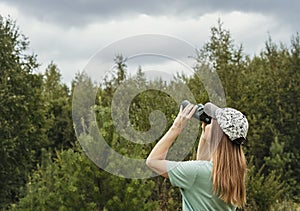 Young blonde woman bird watcher in cap and blue t-shirt looking through binoculars at cloudy sky in summer forest ornithological