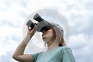 Young blonde woman bird watcher in cap and blue t-shirt looking through binoculars at cloudy sky in summer forest ornithological