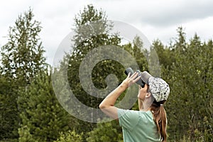 Young blonde woman bird watcher in cap and blue t-shirt looking through binoculars at cloudy sky in summer forest ornithological