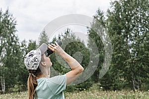 Young blonde woman bird watcher in cap and blue t-shirt looking through binoculars at cloudy sky in summer forest ornithological