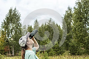 Young blonde woman bird watcher in cap and blue t-shirt looking through binoculars at cloudy sky in summer forest ornithological