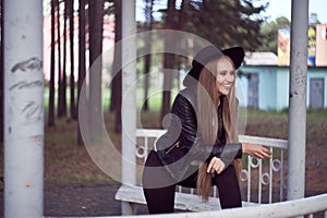 A young blonde woman in a black hat inside an architectural structure. A black leather jacket and a white shirt