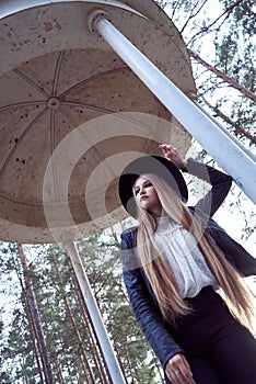 A young blonde woman in a black hat inside an architectural structure. A black leather jacket and a white shirt