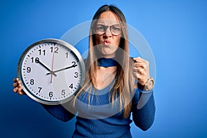Young blonde woman with blue eyes holding big minute clock over isolated background annoyed and frustrated shouting with anger,