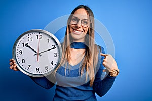 Young blonde woman with blue eyes holding big minute clock over isolated background with surprise face pointing finger to himself