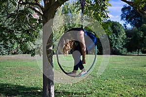 Young blonde woman in a blue suit, exercising on an aero hoop hanging from a tree in the middle of a park. Concept aerial hoop,