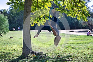 Young blonde woman in a blue suit, exercising upside down in an aero hoop hanging from a tree in the middle of a park. Concept