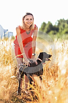 A young blonde woman in a bright dress stands in the middle of a field with a hunting dog. Happiness, freedom, summer, vacation,