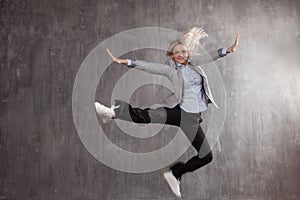 Young blonde woman in business suit and sneakers jumping for joy, gray textured background.