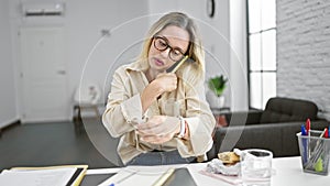 Young blonde woman business worker eating sandwich talking on smartphone at the office