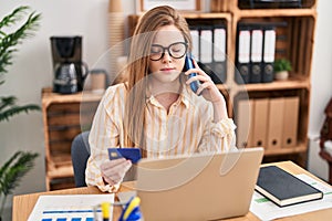 Young blonde woman business worker talking on the smartphone using credit card at office