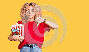 Young blonde woman with curly hair eating popcorn with angry face, negative sign showing dislike with thumbs down, rejection