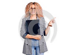 Young blonde woman with curly hair holding business folder smiling happy pointing with hand and finger to the side