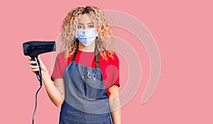 Young blonde woman with curly hair holding dryer blow wearing safety mask for coranvirus looking positive and happy standing and