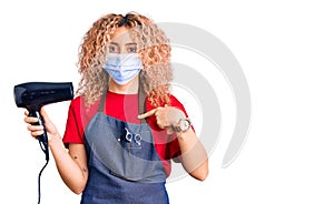 Young blonde woman with curly hair holding dryer blow wearing safety mask for coranvirus pointing finger to one self smiling happy