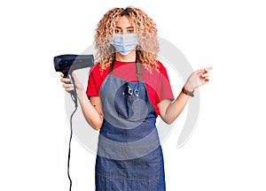 Young blonde woman with curly hair holding dryer blow wearing safety mask for coranvirus smiling happy pointing with hand and