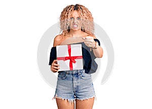 Young blonde woman with curly hair holding gift annoyed and frustrated shouting with anger, yelling crazy with anger and hand