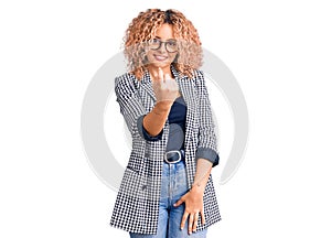 Young blonde woman with curly hair wearing business jacket and glasses beckoning come here gesture with hand inviting welcoming