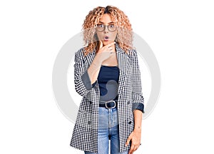 Young blonde woman with curly hair wearing business jacket and glasses looking fascinated with disbelief, surprise and amazed