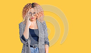 Young blonde woman with curly hair wearing business jacket and glasses pointing to the eye watching you gesture, suspicious