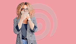 Young blonde woman with curly hair wearing business jacket and glasses shocked covering mouth with hands for mistake