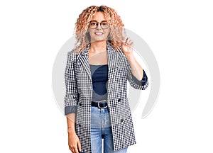 Young blonde woman with curly hair wearing business jacket and glasses showing and pointing up with finger number one while