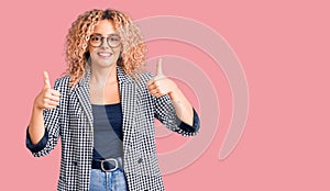 Young blonde woman with curly hair wearing business jacket and glasses success sign doing positive gesture with hand, thumbs up