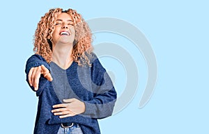 Young blonde woman with curly hair wearing casual winter sweater laughing at you, pointing finger to the camera with hand over