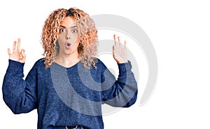 Young blonde woman with curly hair wearing casual winter sweater looking surprised and shocked doing ok approval symbol with