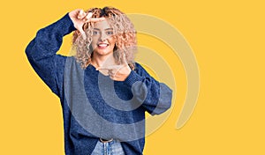 Young blonde woman with curly hair wearing casual winter sweater smiling making frame with hands and fingers with happy face