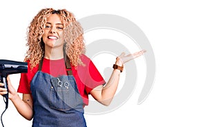 Young blonde woman with curly hair wearing hairdresser apron and holding dryer blow celebrating victory with happy smile and
