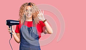 Young blonde woman with curly hair wearing hairdresser apron and holding dryer blow doing ok sign with fingers, smiling friendly