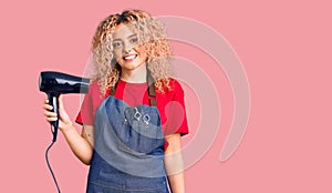 Young blonde woman with curly hair wearing hairdresser apron and holding dryer blow looking positive and happy standing and