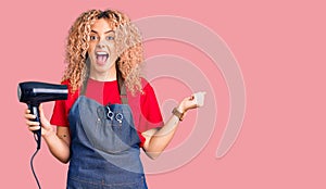 Young blonde woman with curly hair wearing hairdresser apron and holding dryer blow screaming proud, celebrating victory and