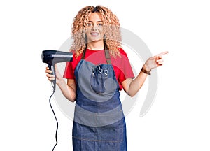 Young blonde woman with curly hair wearing hairdresser apron and holding dryer blow smiling happy pointing with hand and finger to