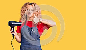 Young blonde woman with curly hair wearing hairdresser apron and holding dryer blow smiling happy and positive, thumb up doing