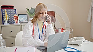 Young blonde woman doctor using laptop talking on smartphone at clinic