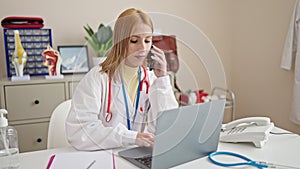 Young blonde woman doctor using laptop talking on smartphone at clinic